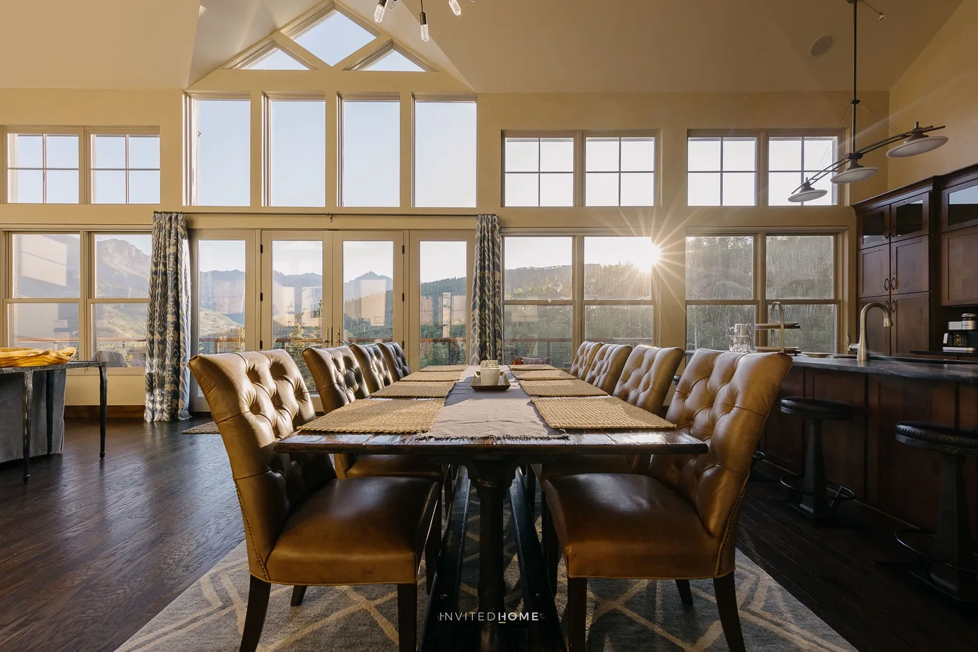 A dining room with a long table and cushioned seating beneath tall windows.