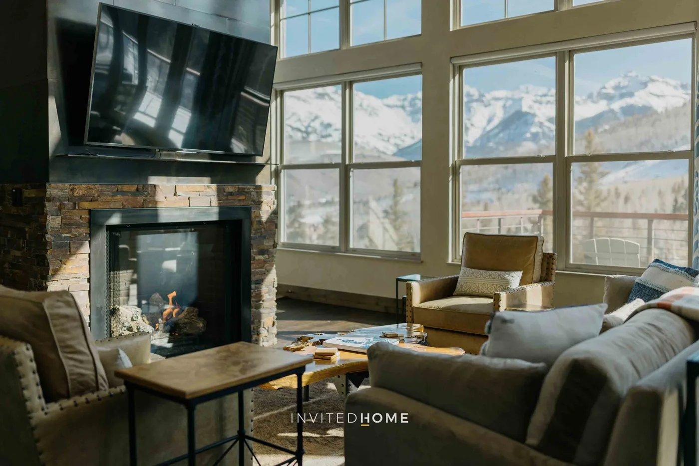 A living room with seating around a fireplace and large windows overlooking snowy mountains.