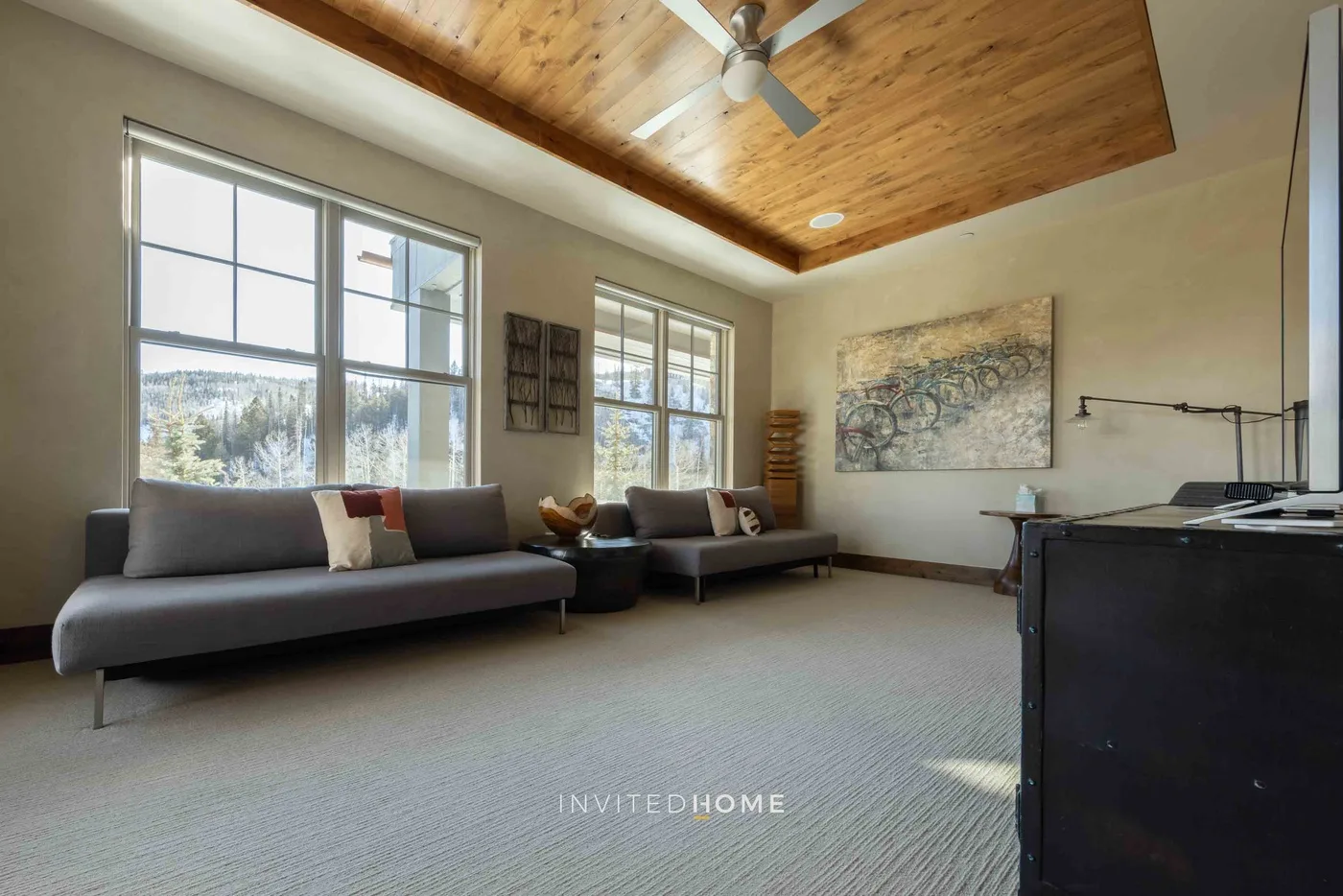 A bright living room with seating beneath a wood-paneled ceiling and large windows.