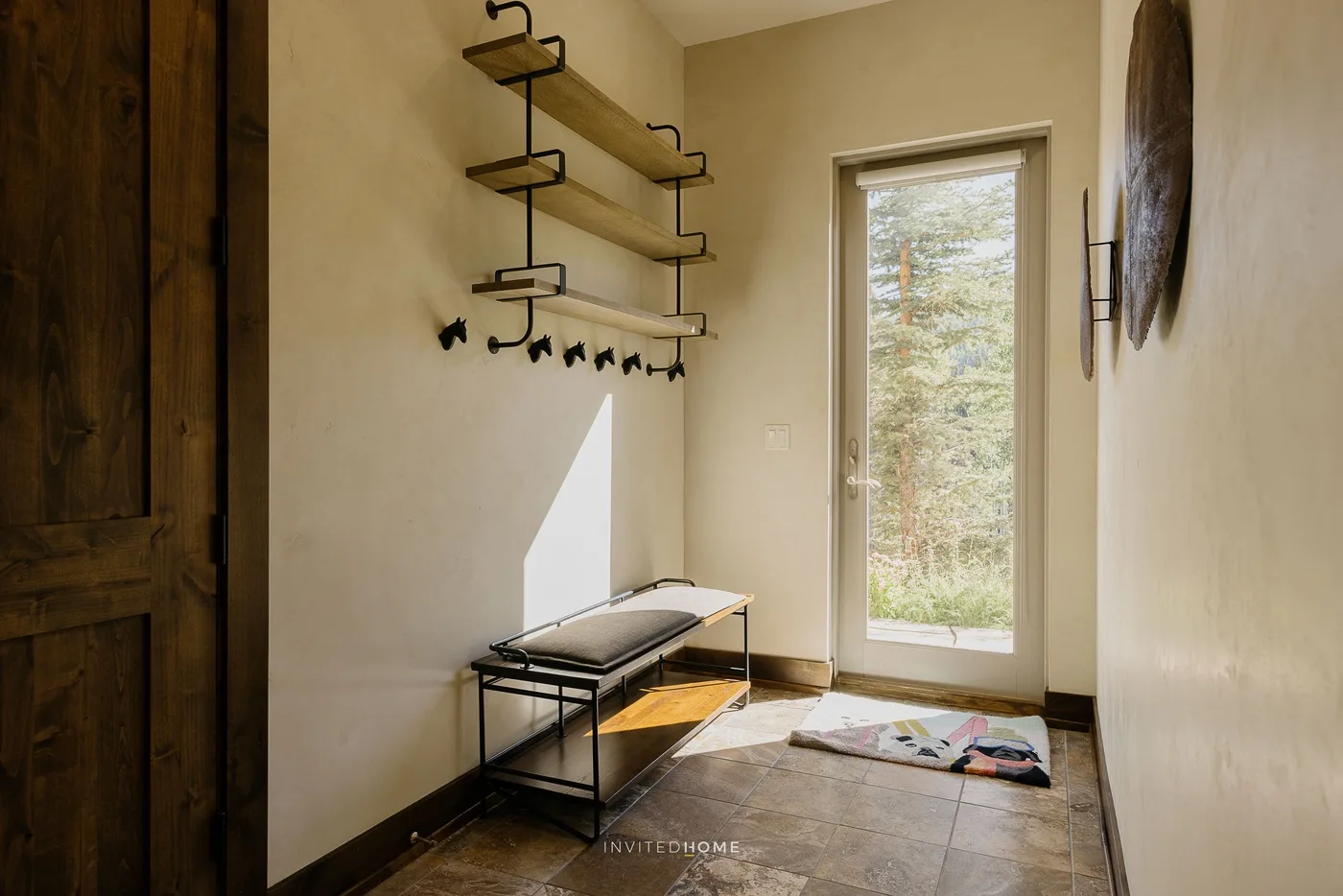 A bright entryway with a bench, wall hooks, and a glass door overlooking trees.
