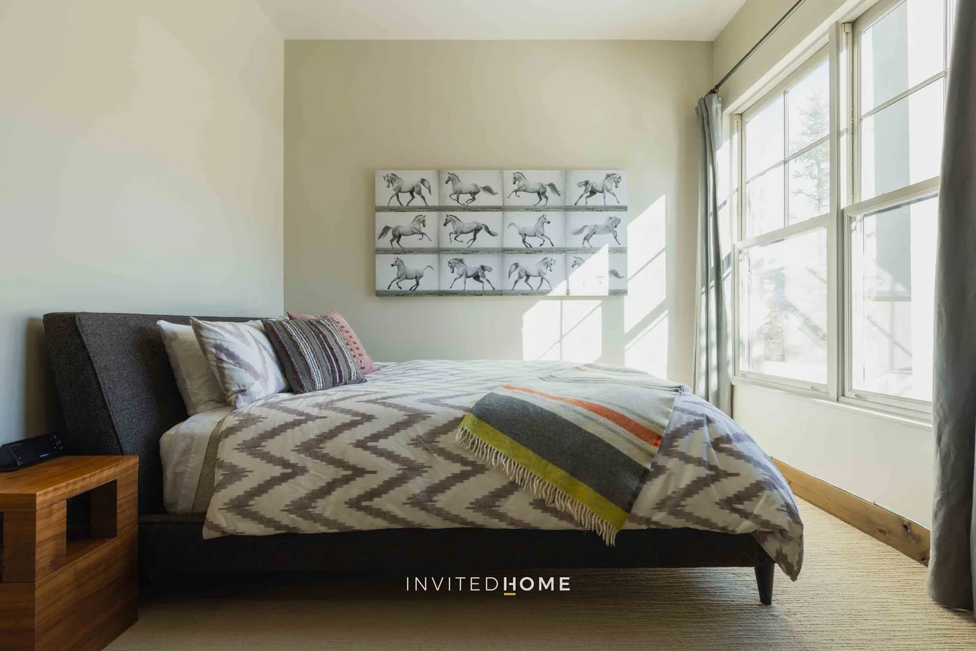 A bright bedroom with a bed beside large windows and horse artwork on the wall.