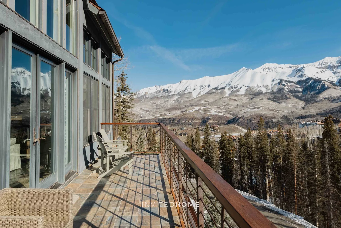 A balcony with outdoor seating overlooks a forested valley and snow-covered mountains.