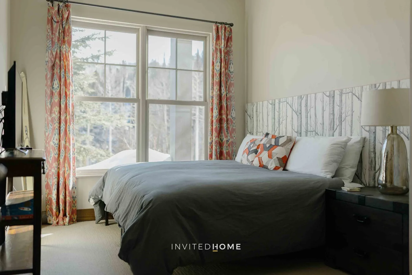 A bedroom with a bed beside a large window and patterned curtains.