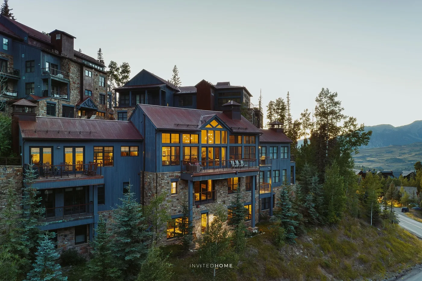A large blue lodge with glowing windows sits among trees beside a mountain road.
