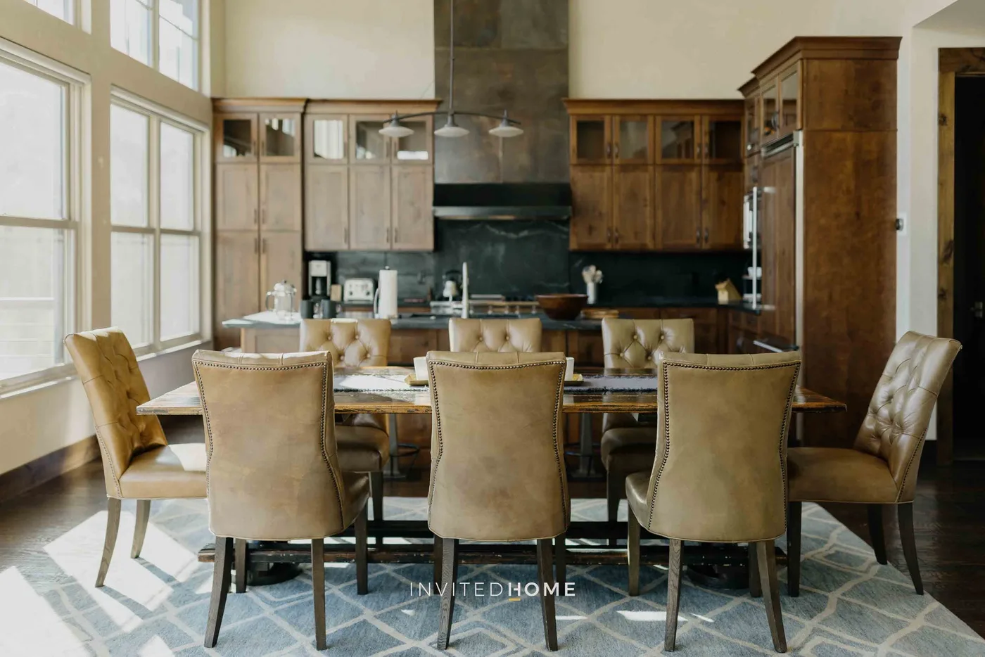 A dining room with a long table and upholstered seating beside tall windows.