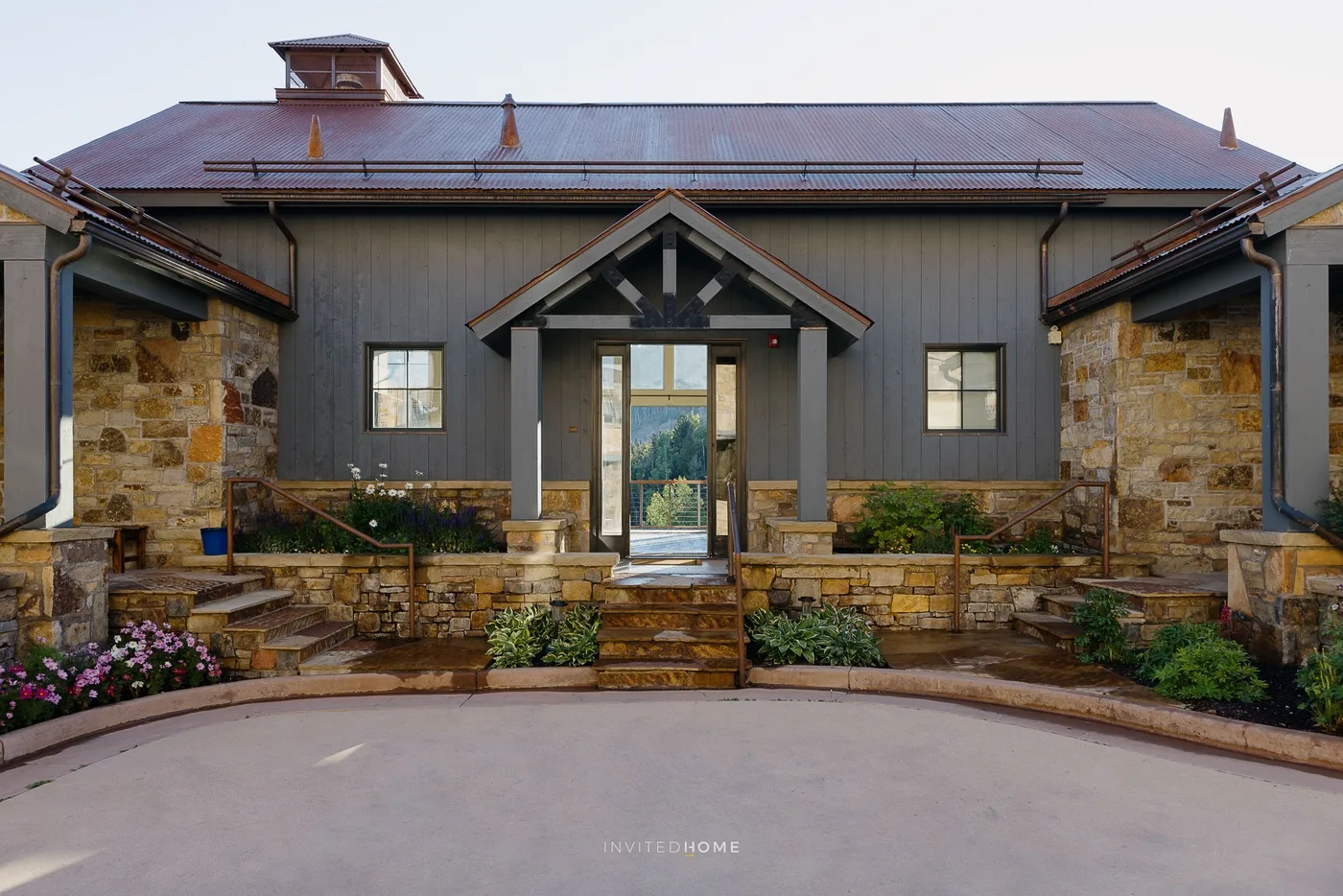 A building entrance with a peaked wooden canopy, stone walls, and landscaped flower beds.