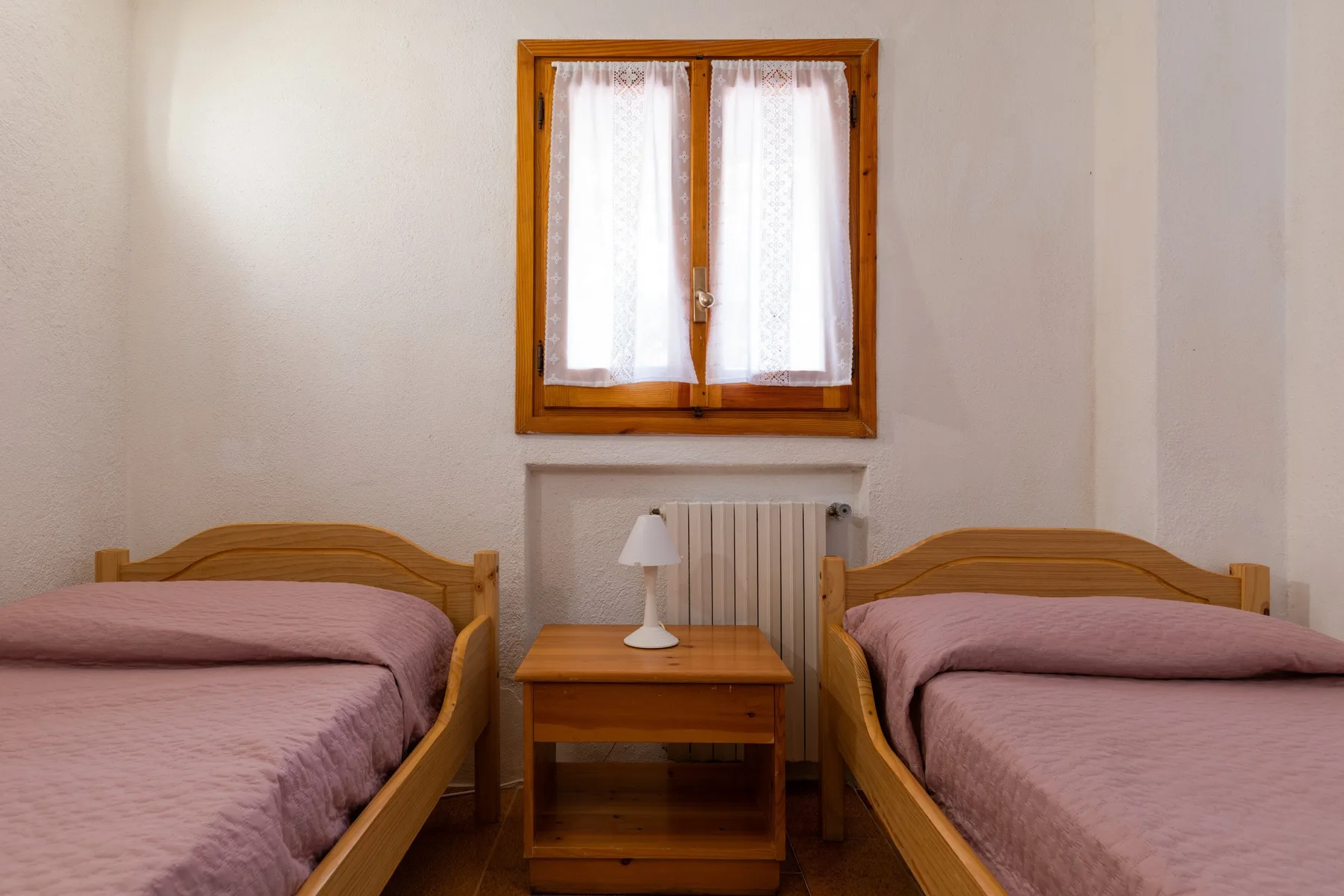 A bedroom with two beds and a small table beneath a curtained window.