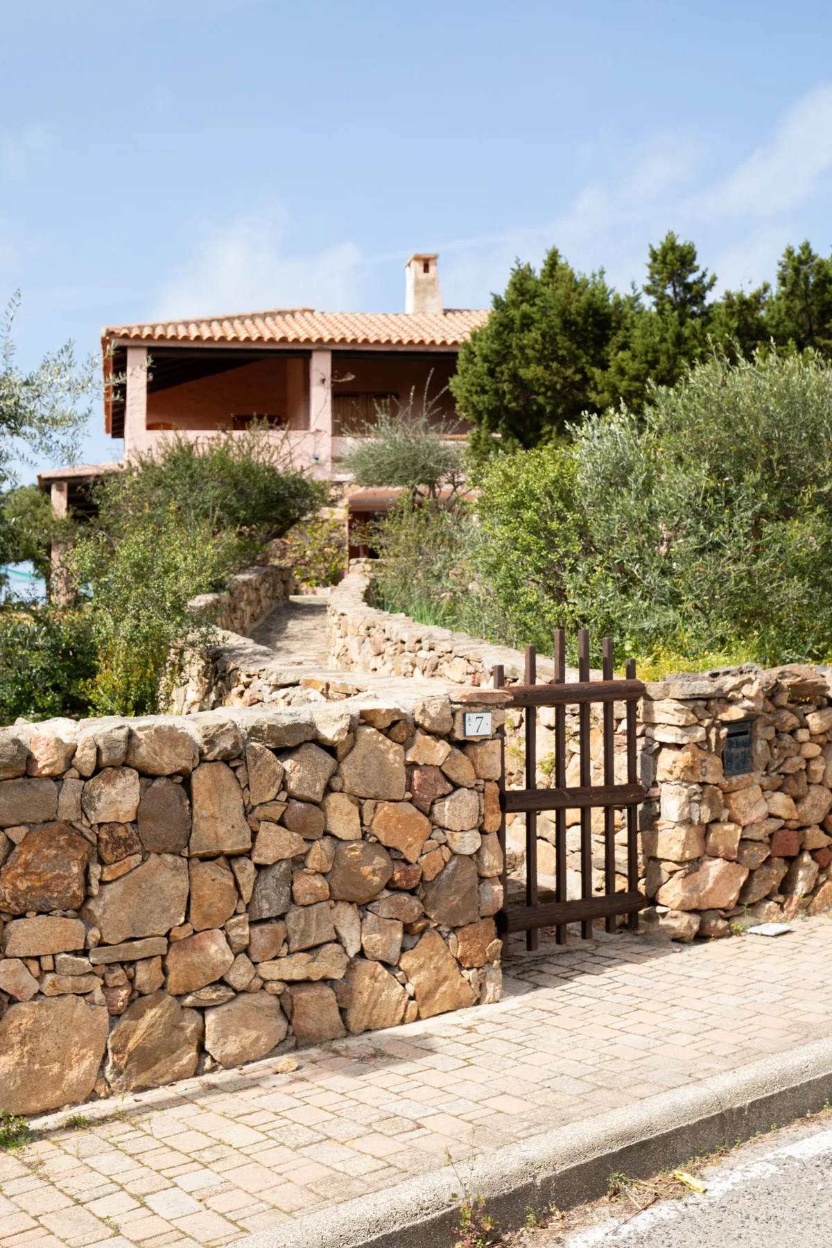 A stone wall and gate border a garden path leading to a house.