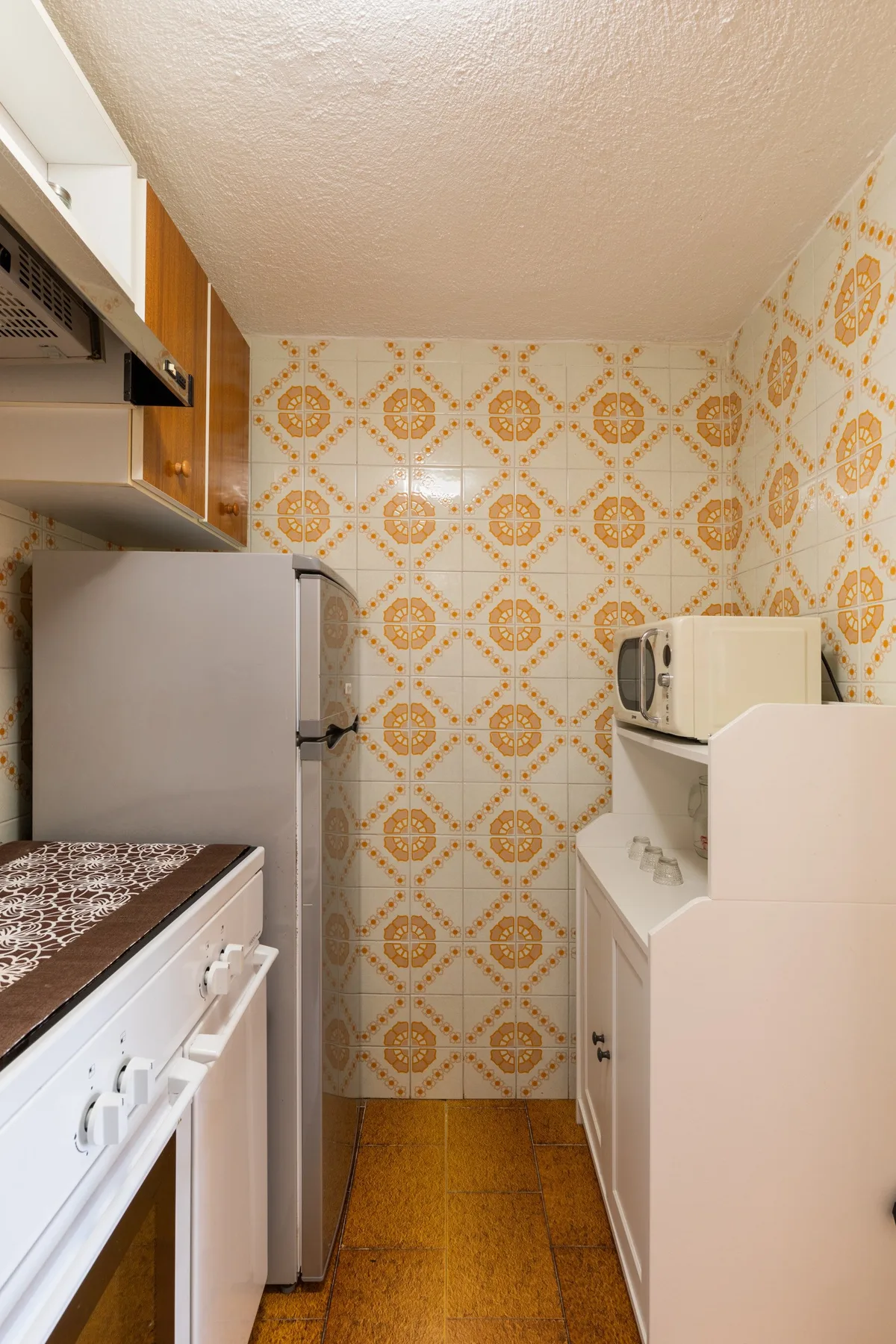 A narrow kitchen with patterned tile walls and appliances along both sides.
