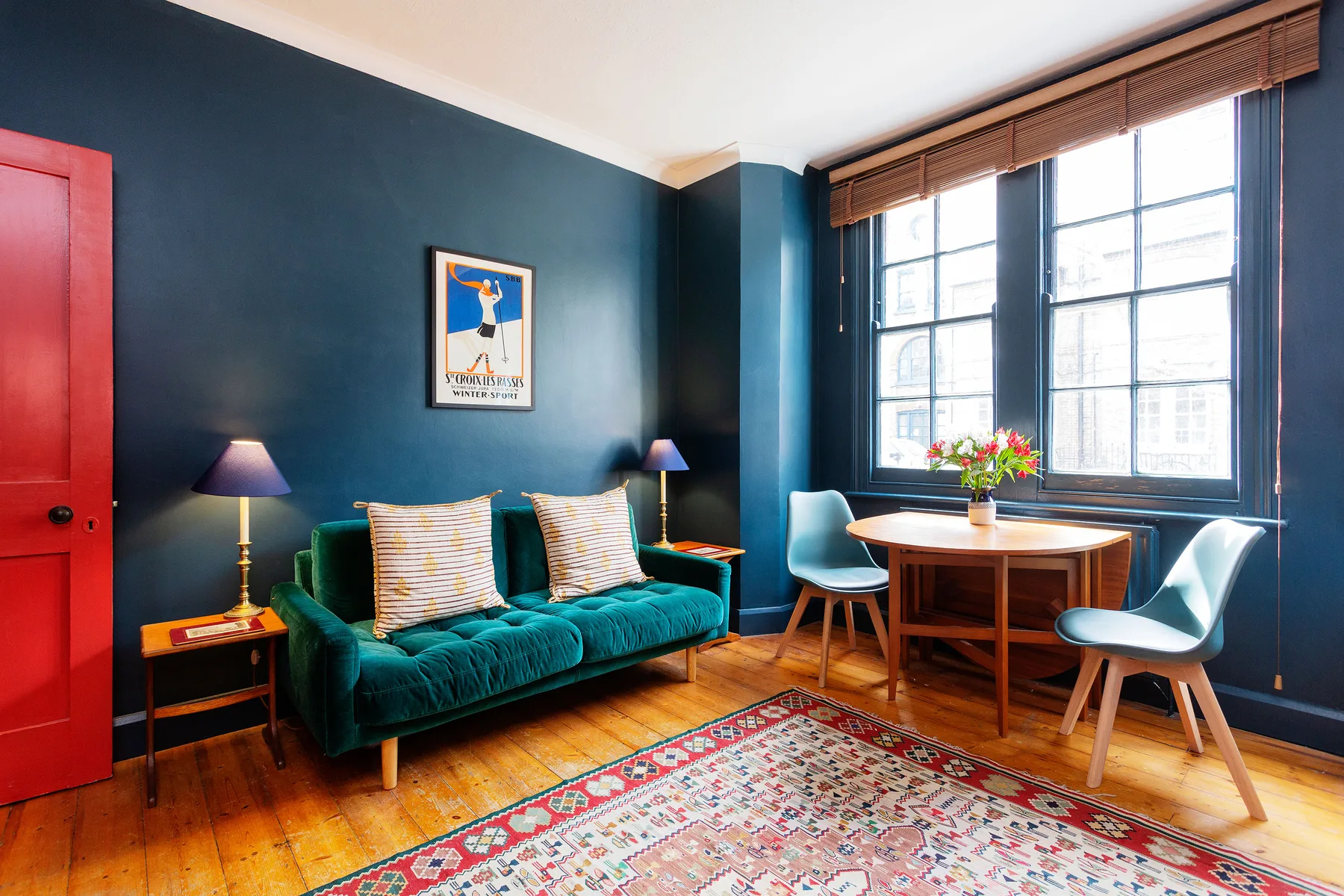 Una luminosa sala de estar tiene un sofá verde azulado junto a una mesa de madera y grandes ventanas.