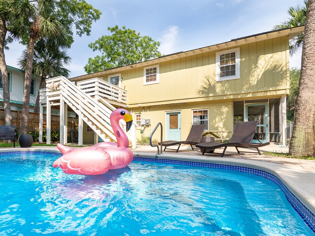 A swimming pool sits beside a yellow two-story building with outdoor seating.