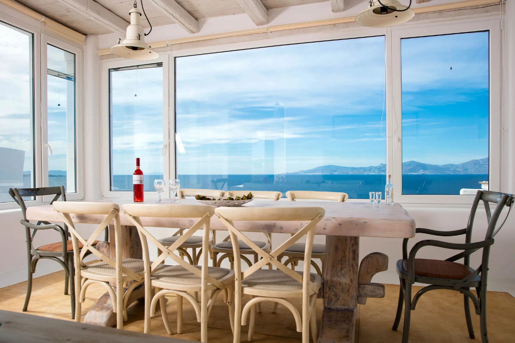 A dining area with a long table beside large windows overlooking the sea.