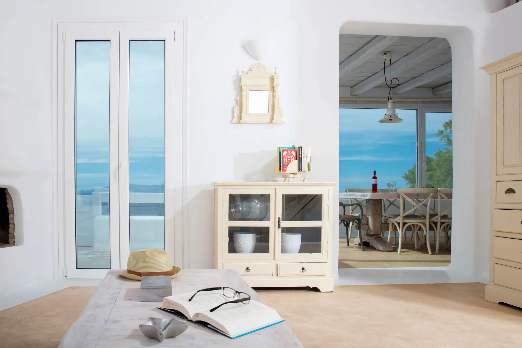 A bright living room opens onto a dining area with blue water visible outside.