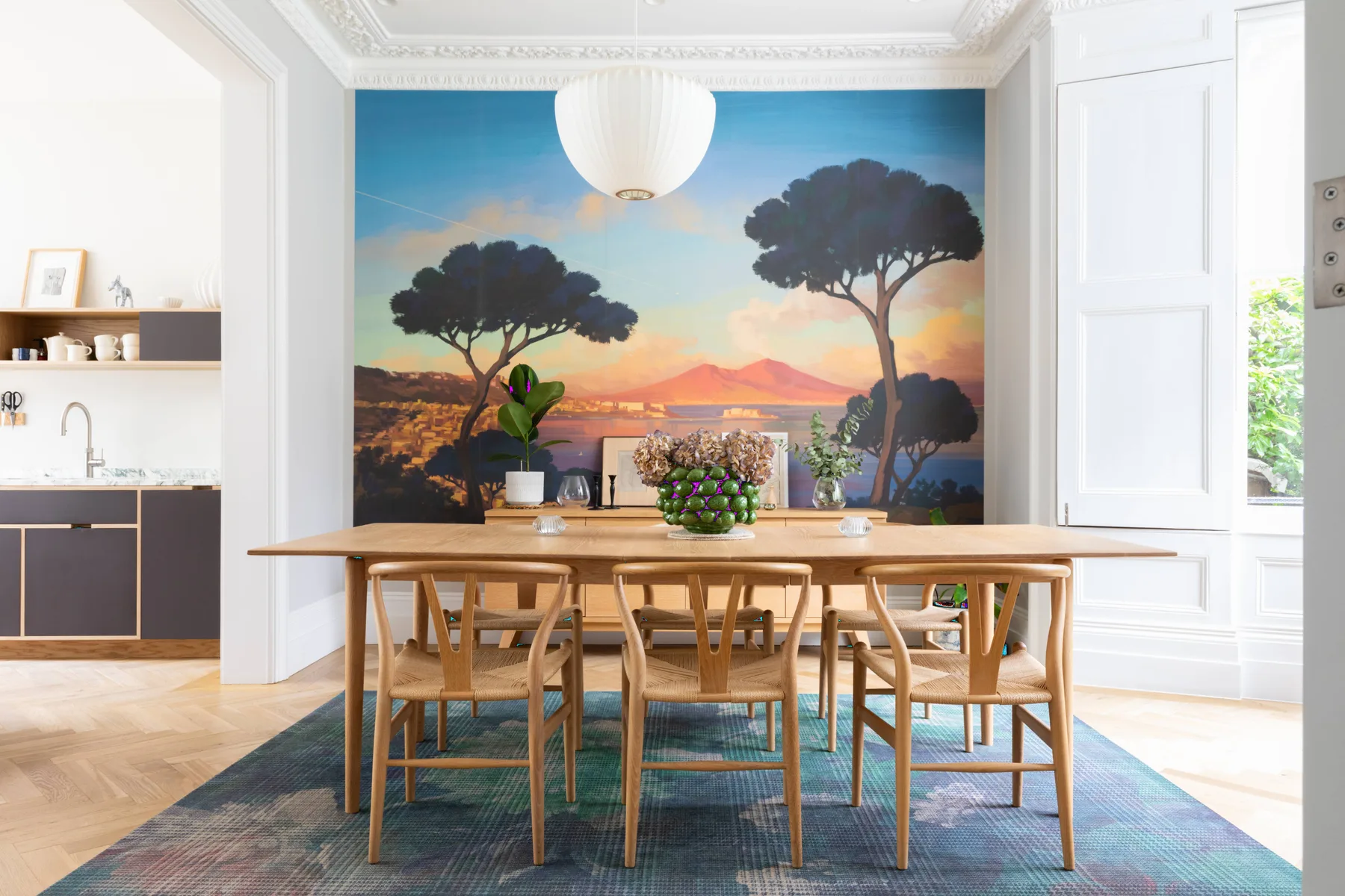 A dining area with a wooden table set before a colorful landscape mural.