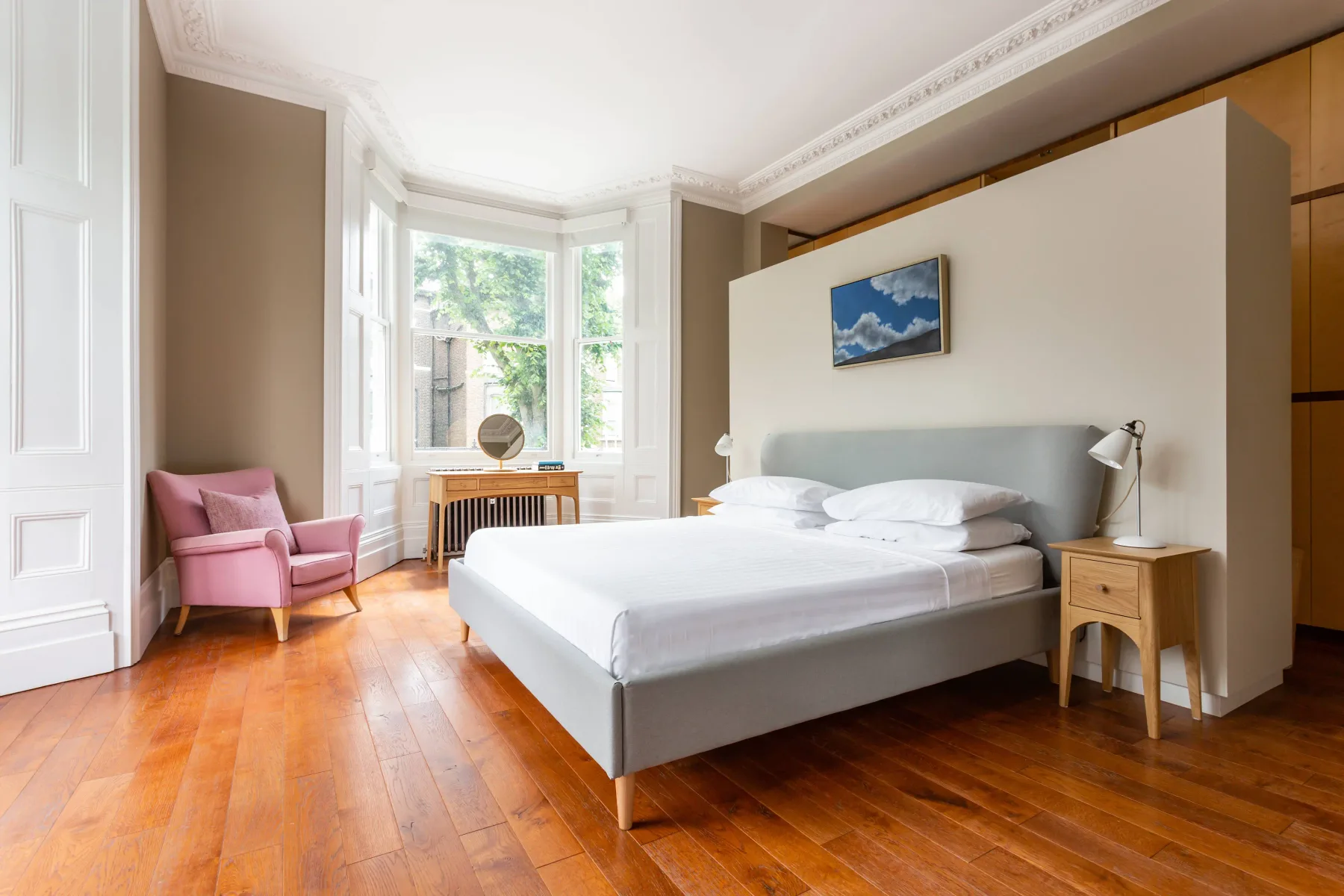 A bright bedroom with a bed, pink seating, and tall bay windows.