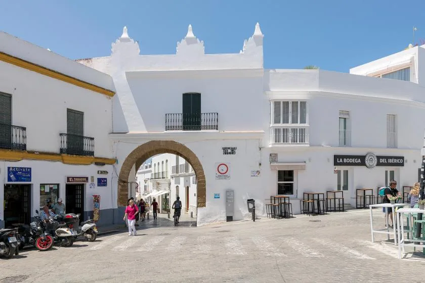 A sunny plaza with white buildings, an arched passage, and outdoor seating.