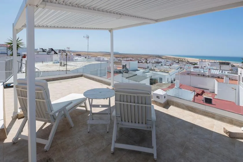 A rooftop terrace with outdoor seating overlooks low-rise buildings and the sea.