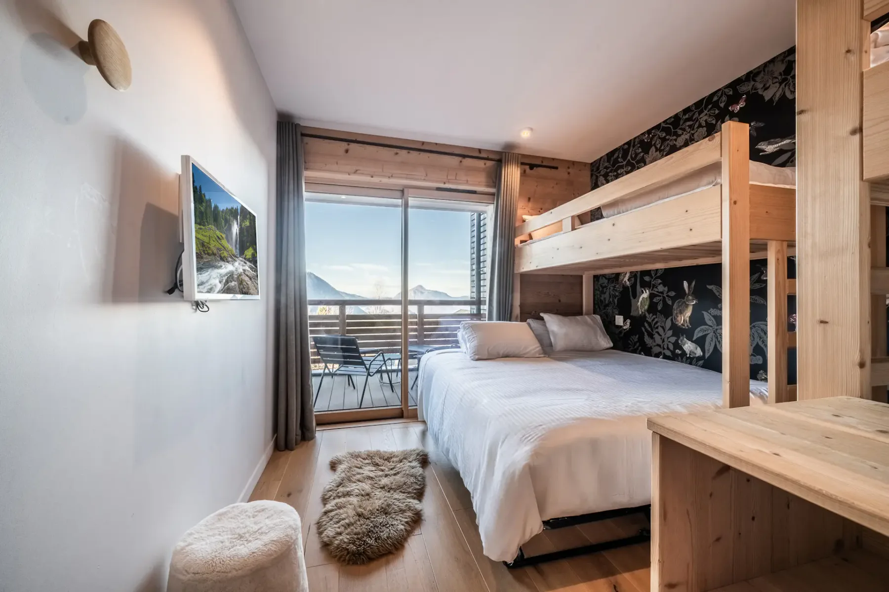 A cozy bedroom features a double bed with a bunk above, a wall-mounted TV, wood accents, and a sliding glass door opening to a balcony with mountain views.
