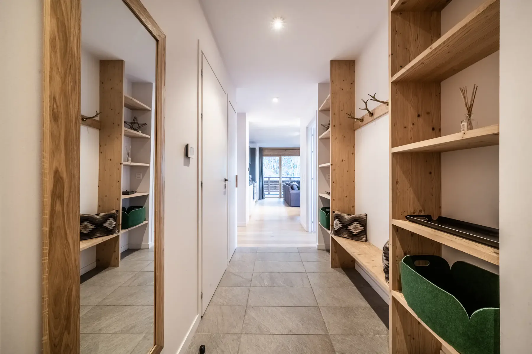 A hallway with wooden shelves, a coat rack, and a mirror. The floor is tiled, and the walls are white.