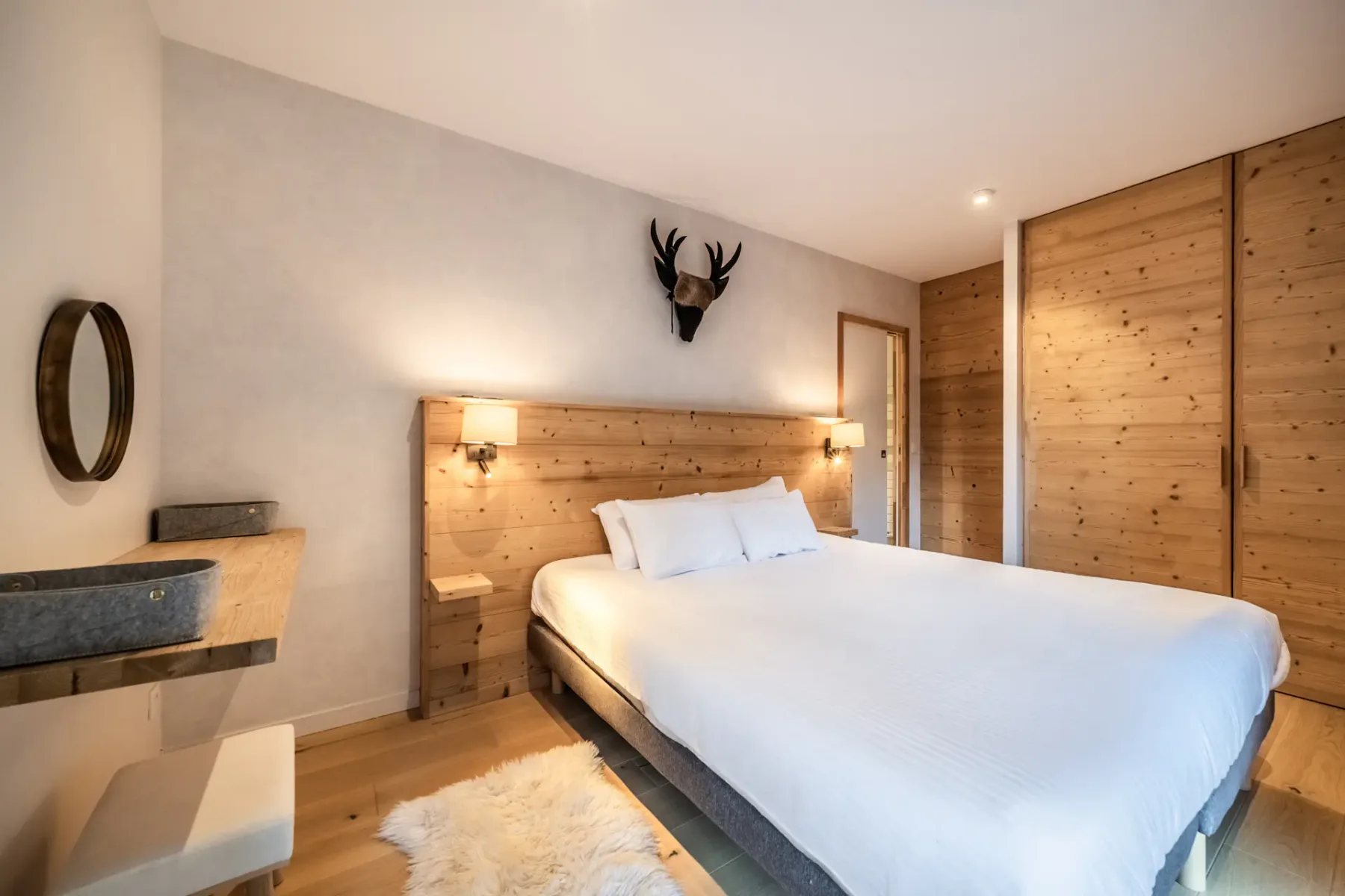 Cozy bedroom with a wooden headboard, two wall lamps, and a decorative deer head above the bed. The room features white bedding, a round mirror, and a wooden floor. A door is open to another room.