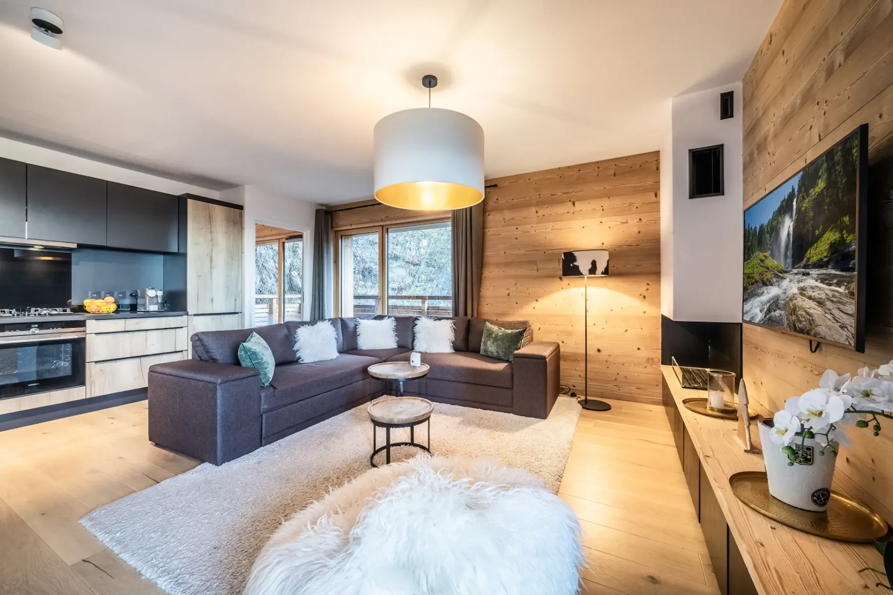 Modern living room with a large brown sectional sofa, wood-paneled walls, and a flat-screen TV. The room features a round chandelier, fluffy white rug, and a view of trees through glass doors.