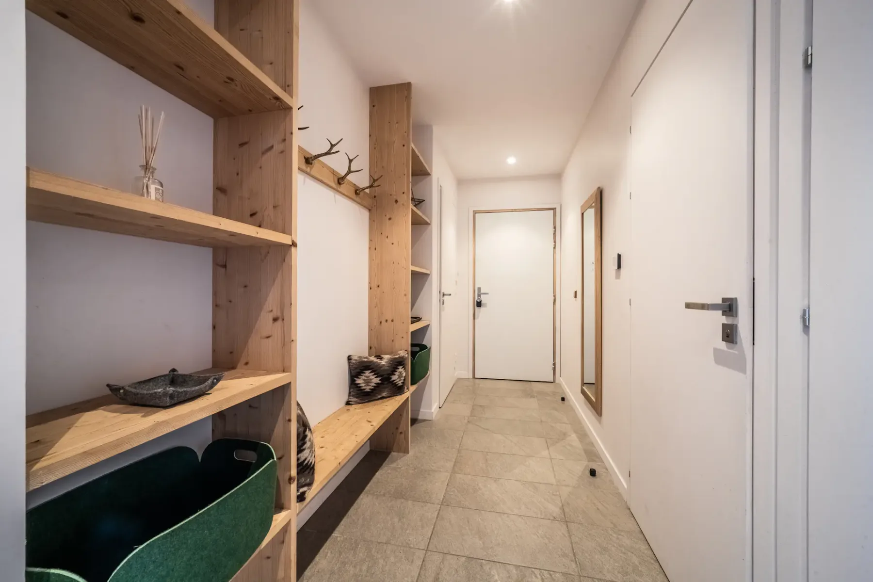 A hallway with wooden shelves, a coat rack, and a mirror. The floor is tiled, and the walls are white.