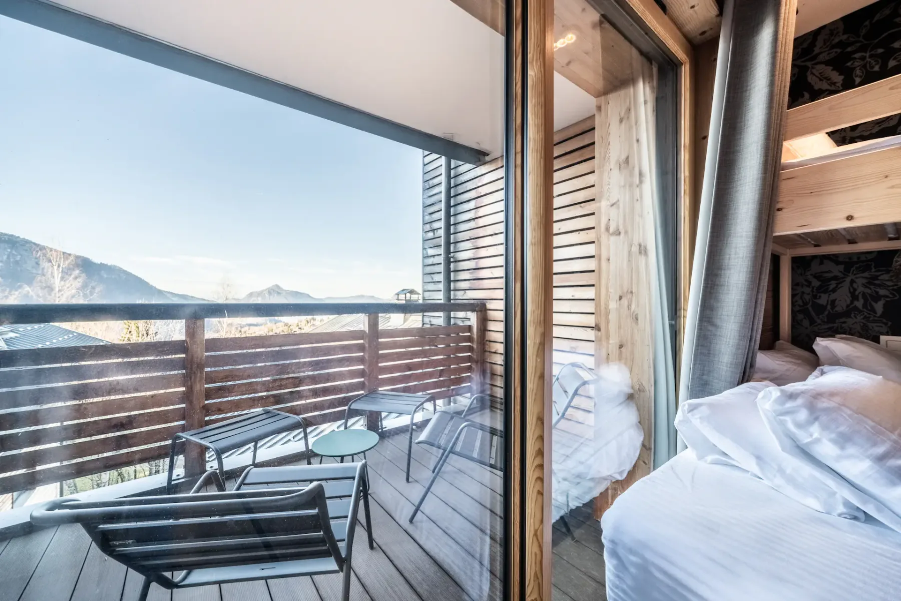 A cozy bedroom features a double bed with a bunk above, a wall-mounted TV, wood accents, and a sliding glass door opening to a balcony with mountain views.