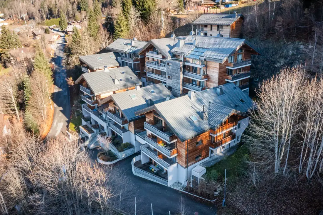 Aerial view of a large multi-story wooden chalet complex surrounded by trees in a mountainous area.