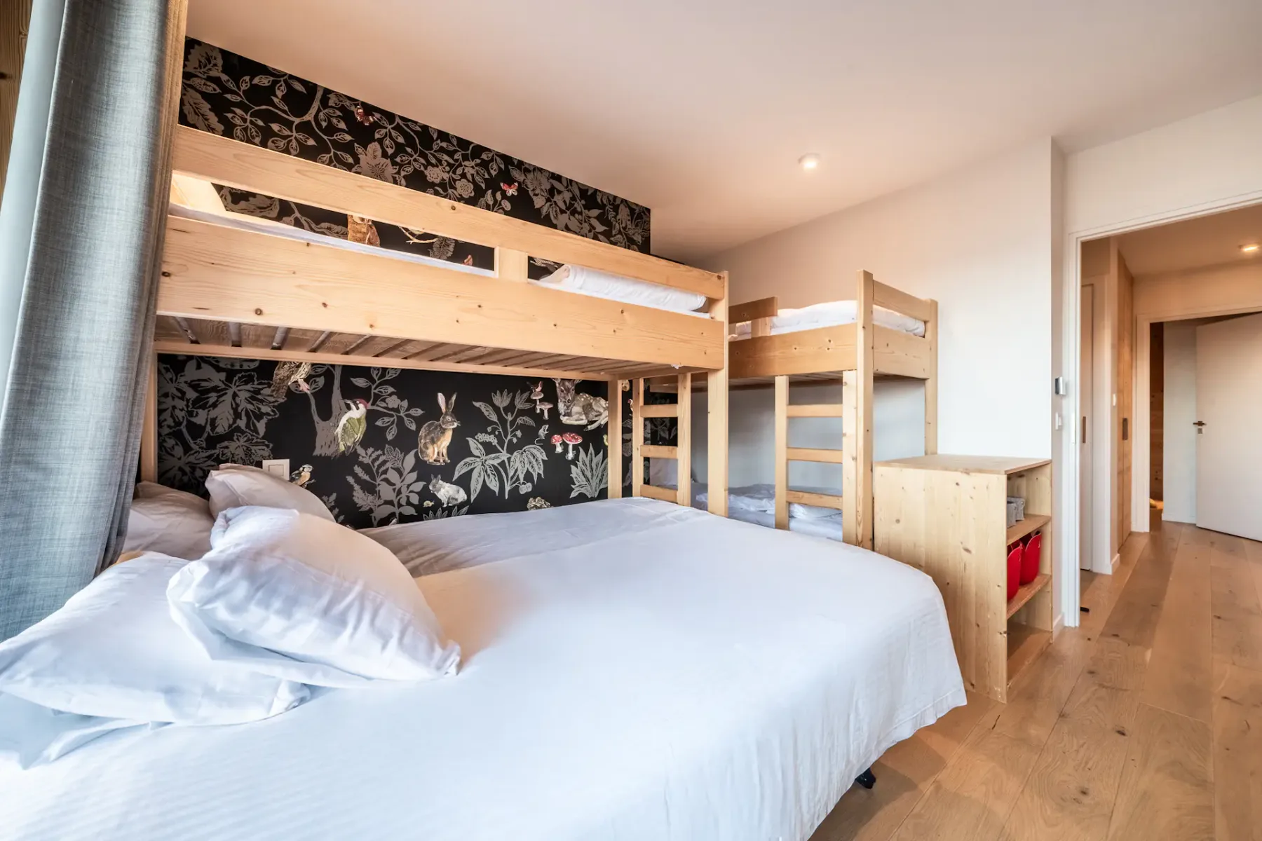 A bedroom with a double bed beneath a wooden bunk bed and floral-patterned wallpaper.