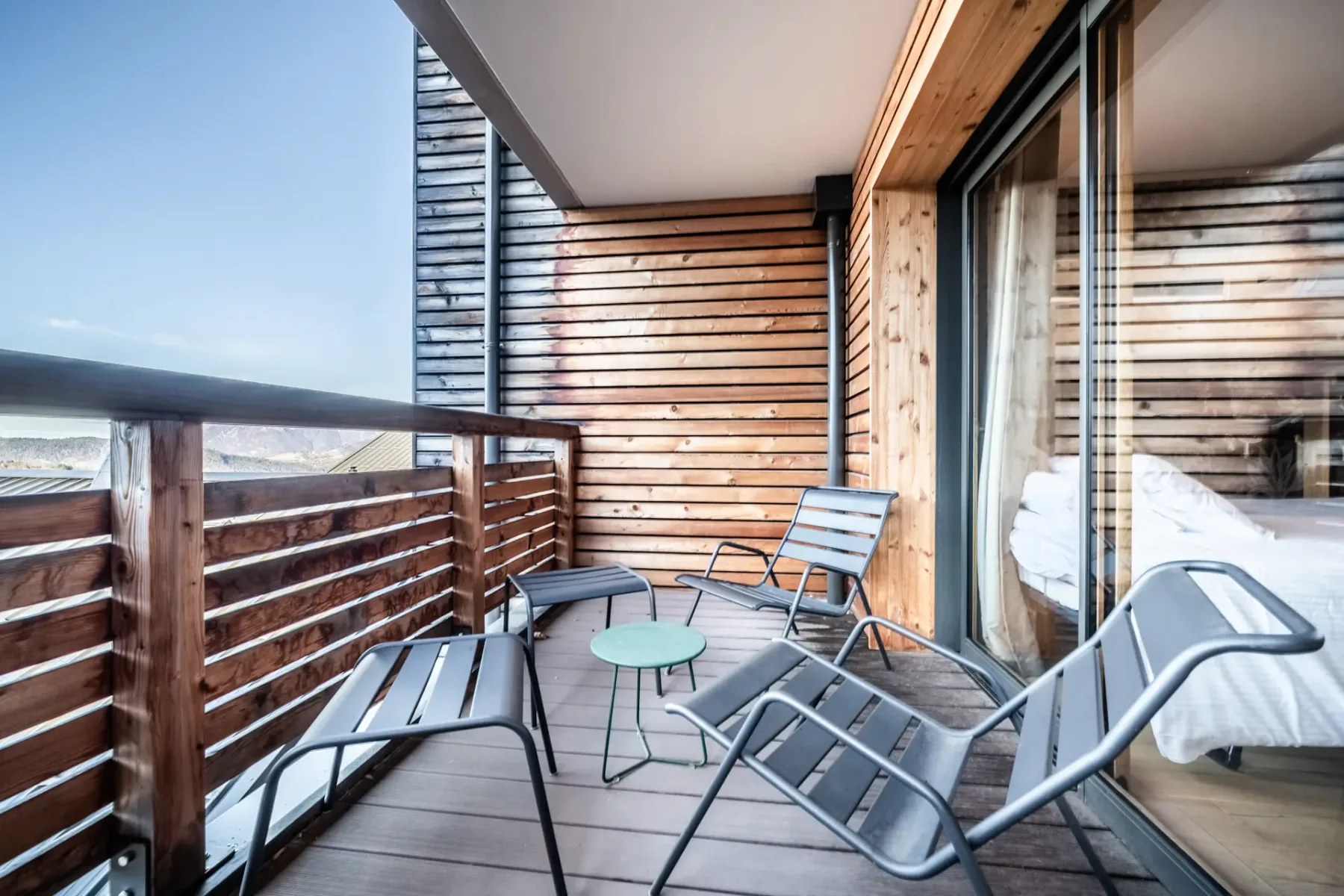 A wooden balcony has outdoor seating and a small round table beside sliding glass doors.