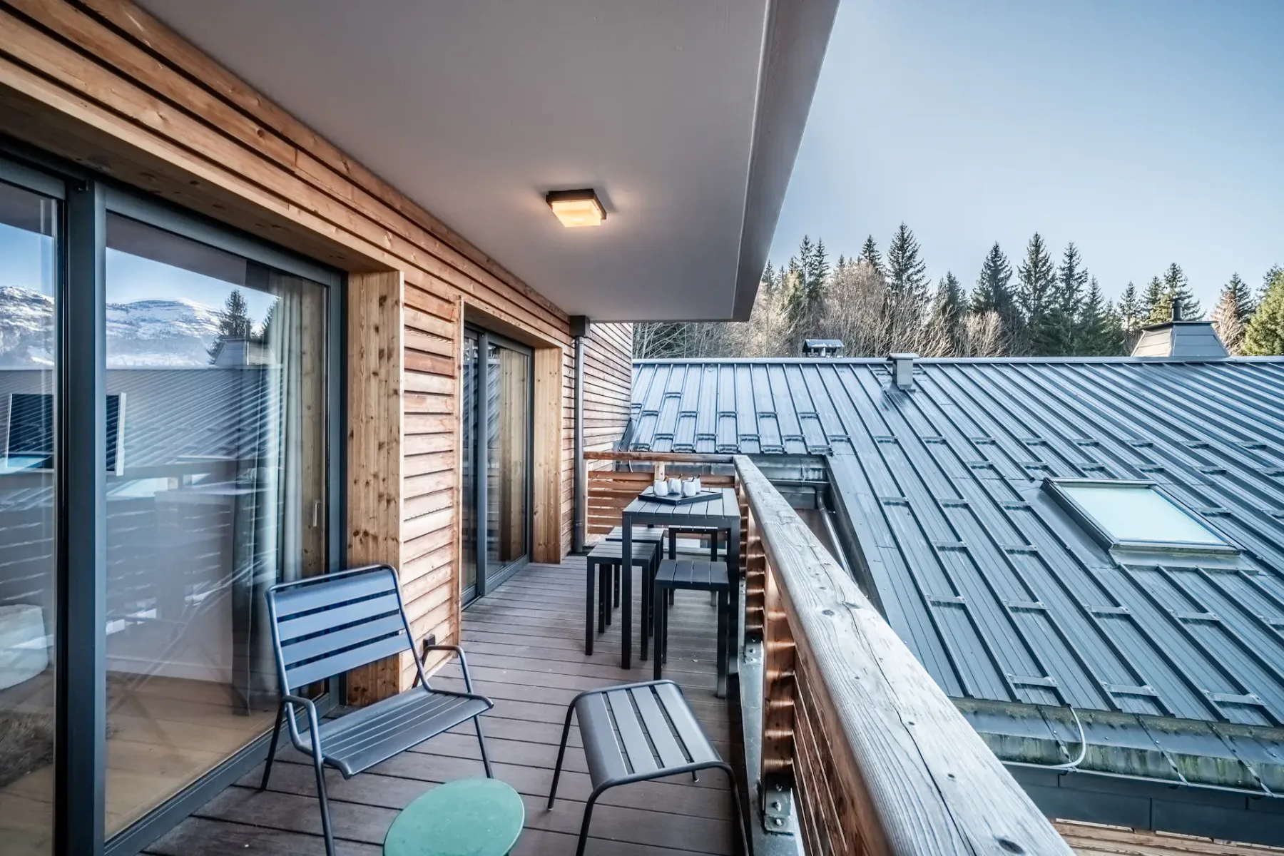 A wooden balcony has outdoor seating beside a metal roof and trees.