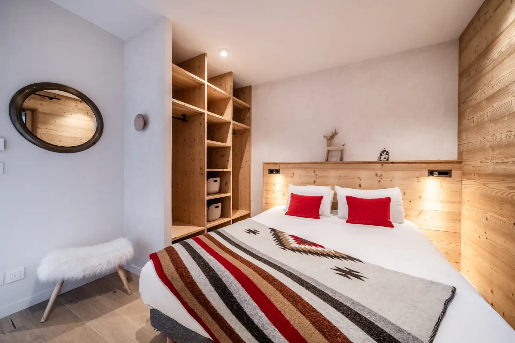 A cozy bedroom with a double bed, red and white pillows, a patterned blanket, wood paneling, open shelving, a circular mirror, and a fluffy bench.