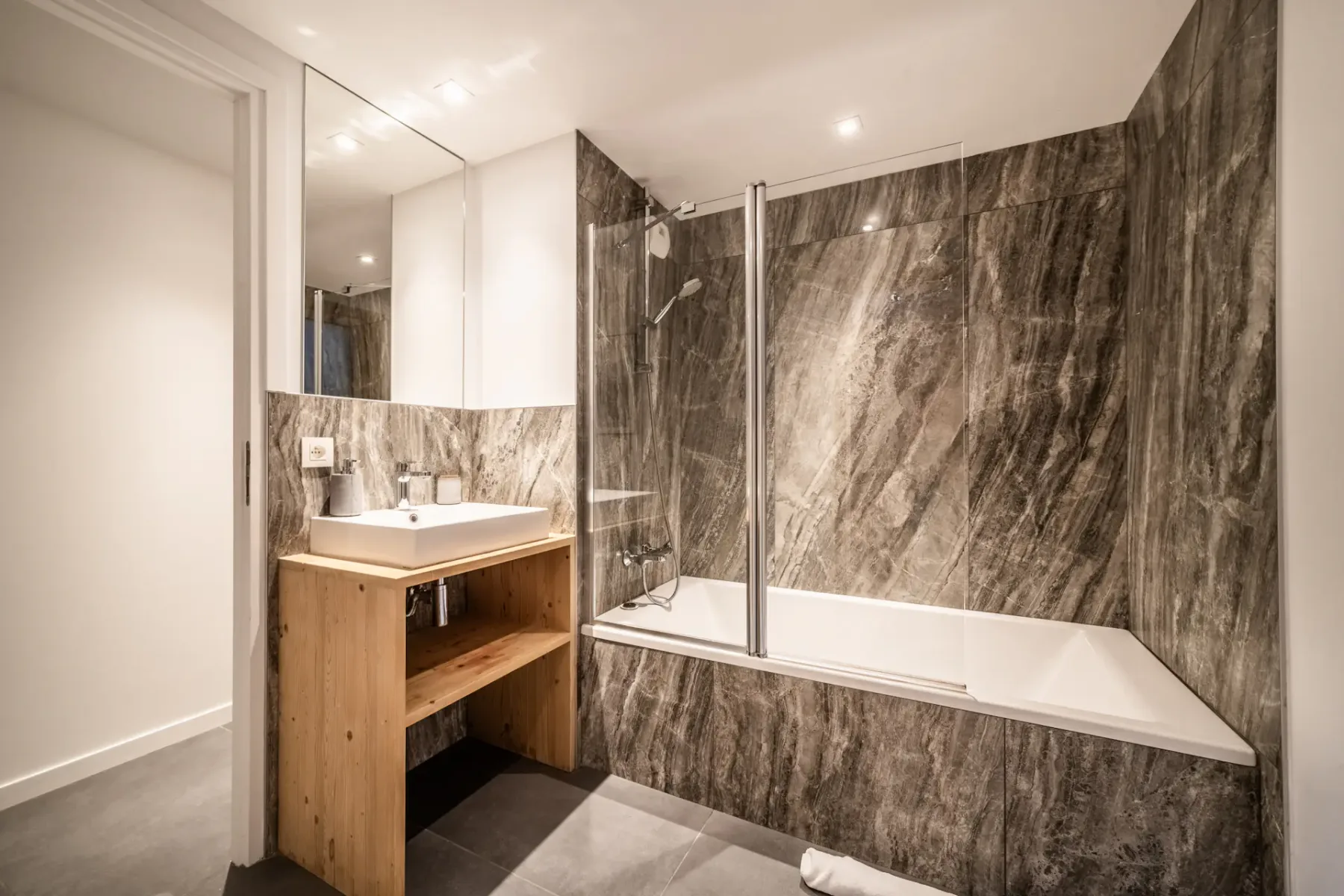 A bathroom with a wooden vanity, wall mirror, and bathtub beside a glass shower screen.