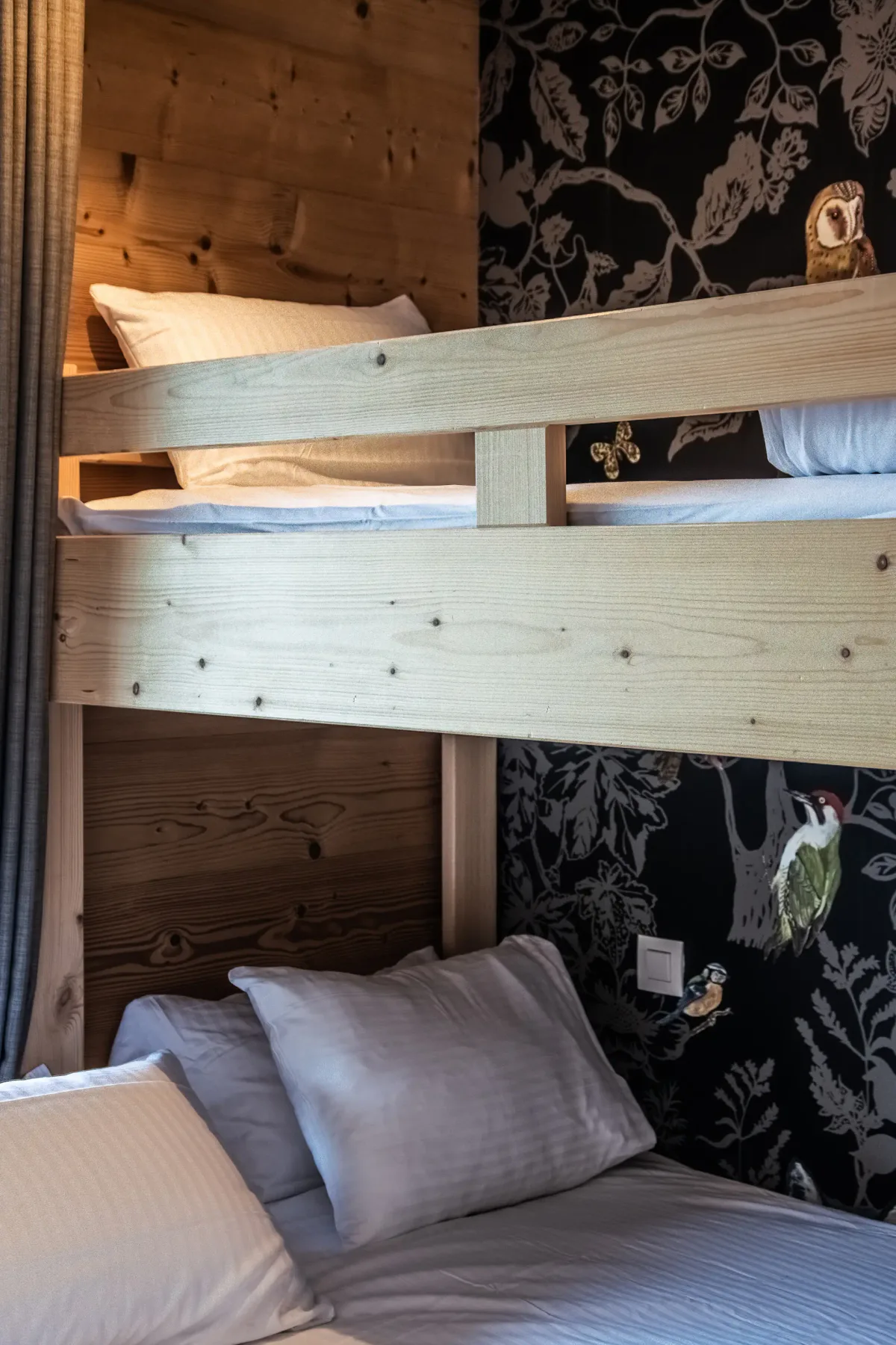 A bedroom with a lower bed beneath an upper bunk and patterned wallpaper.