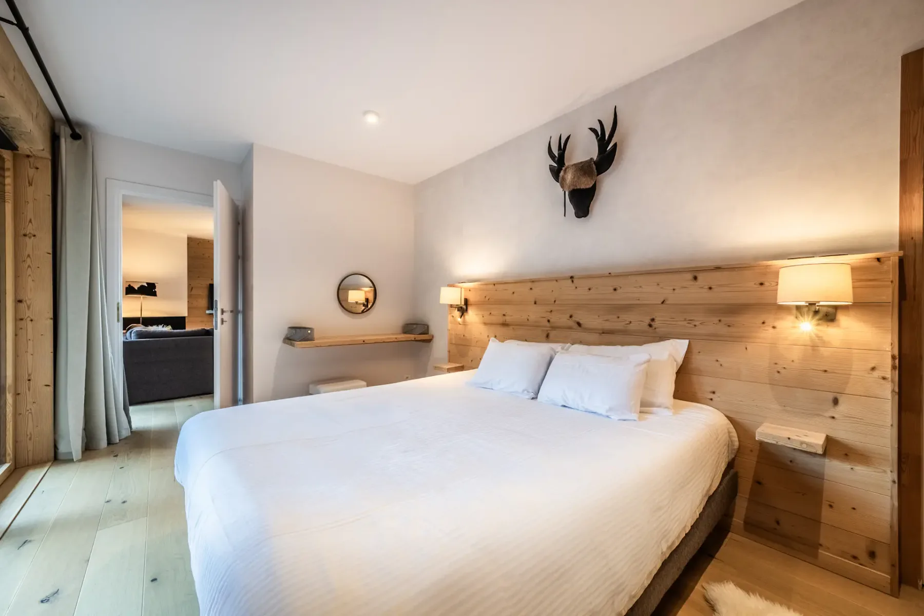 Cozy bedroom with a wooden headboard, two wall lamps, and a decorative deer head above the bed. The room features white bedding, a round mirror, and a wooden floor. A door is open to another room.
