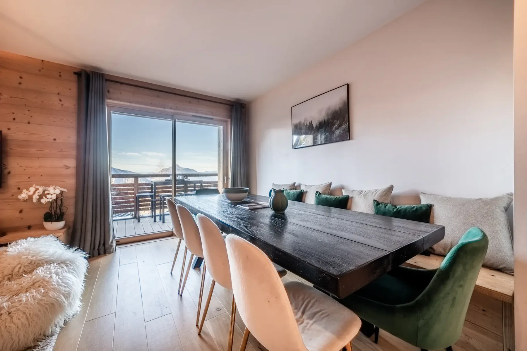 Dining area with a long wooden table, assorted chairs, and a view of a balcony through glass doors. Neutral tones with outdoor scenery visible in the background.