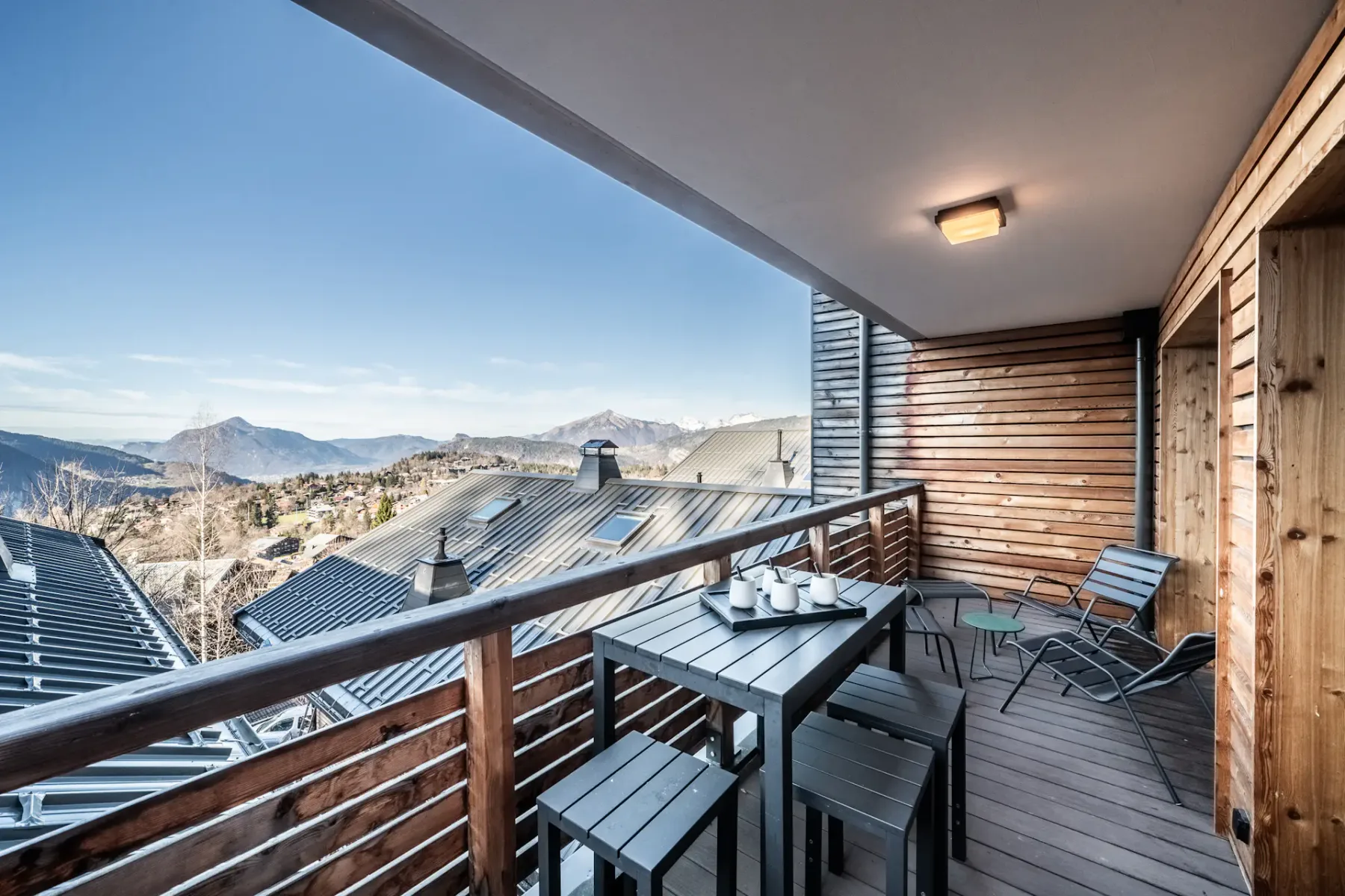 A wooden balcony with a table, cups, and chairs overlooks a scenic mountain view under a clear sky.