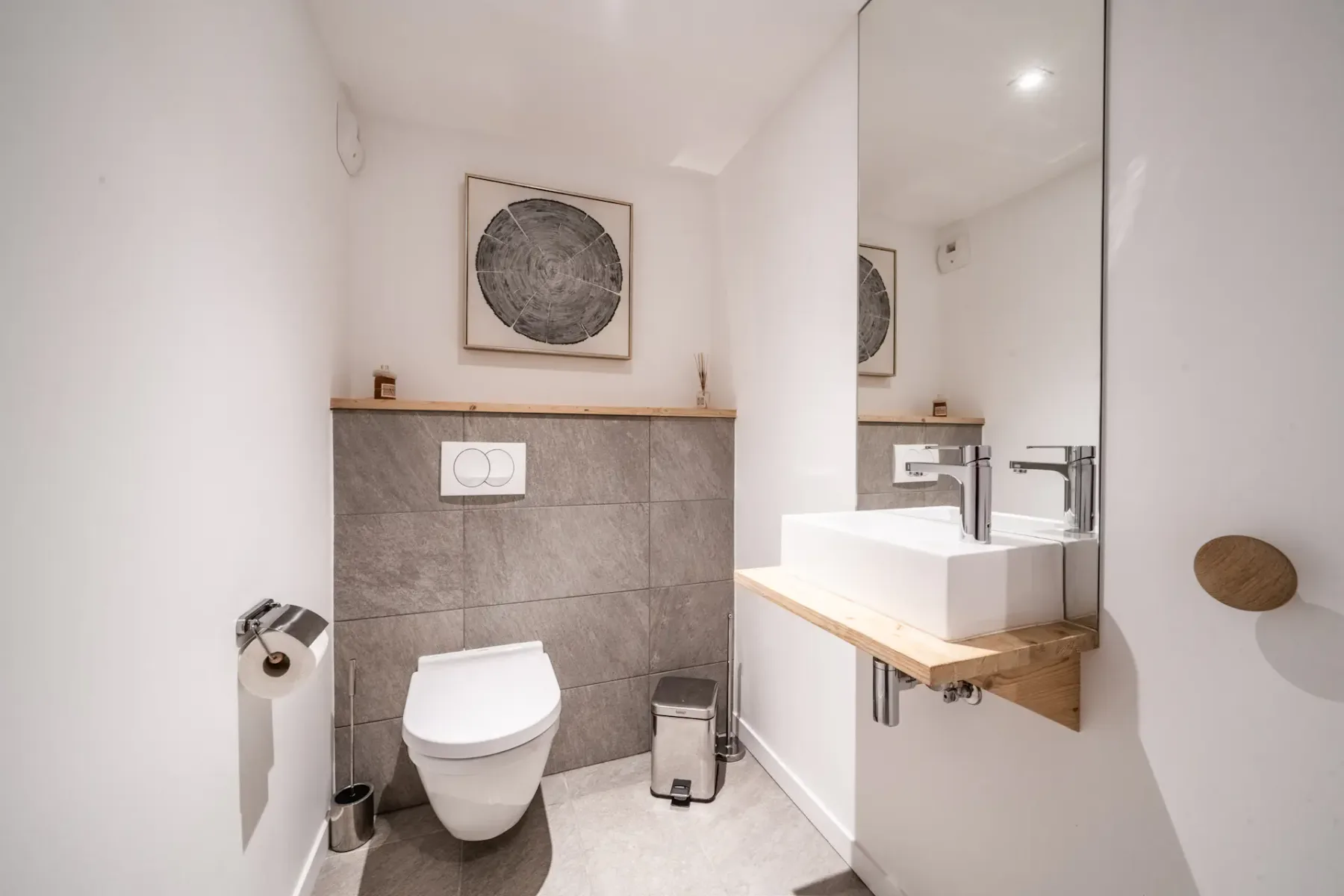 Minimalist bathroom with a wall-mounted toilet, rectangular sink on a wooden counter, large mirror, and framed artwork above gray tiles. Toilet paper holder and waste bin are near the toilet.