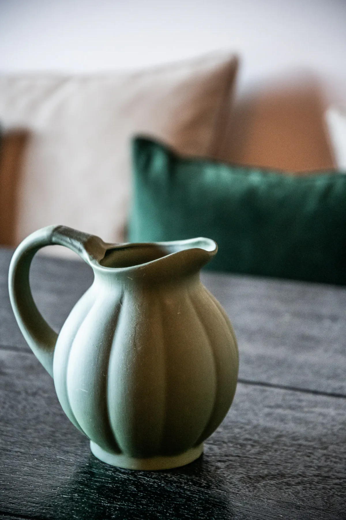 A ceramic pitcher sits on a table in front of cushions.