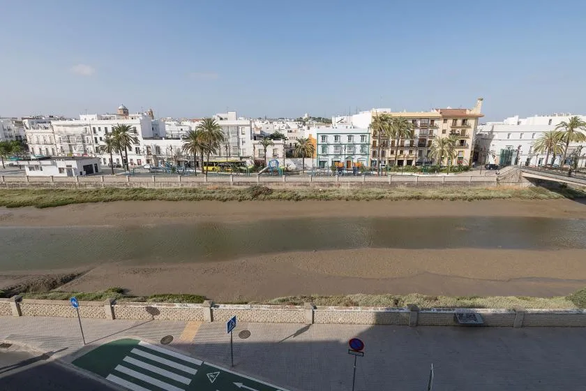 A broad river channel runs past a row of low-rise buildings and palm trees.