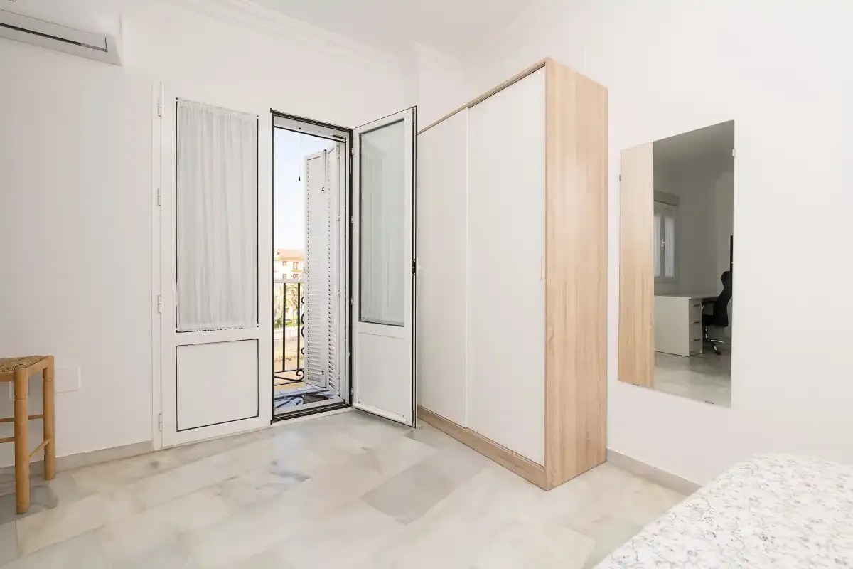 A bright bedroom with an open balcony door and a mirrored wardrobe.