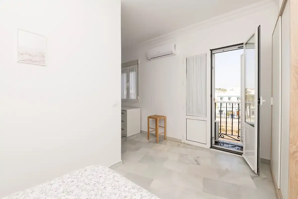 A bright bedroom with a bed, a small table, and an open balcony door.