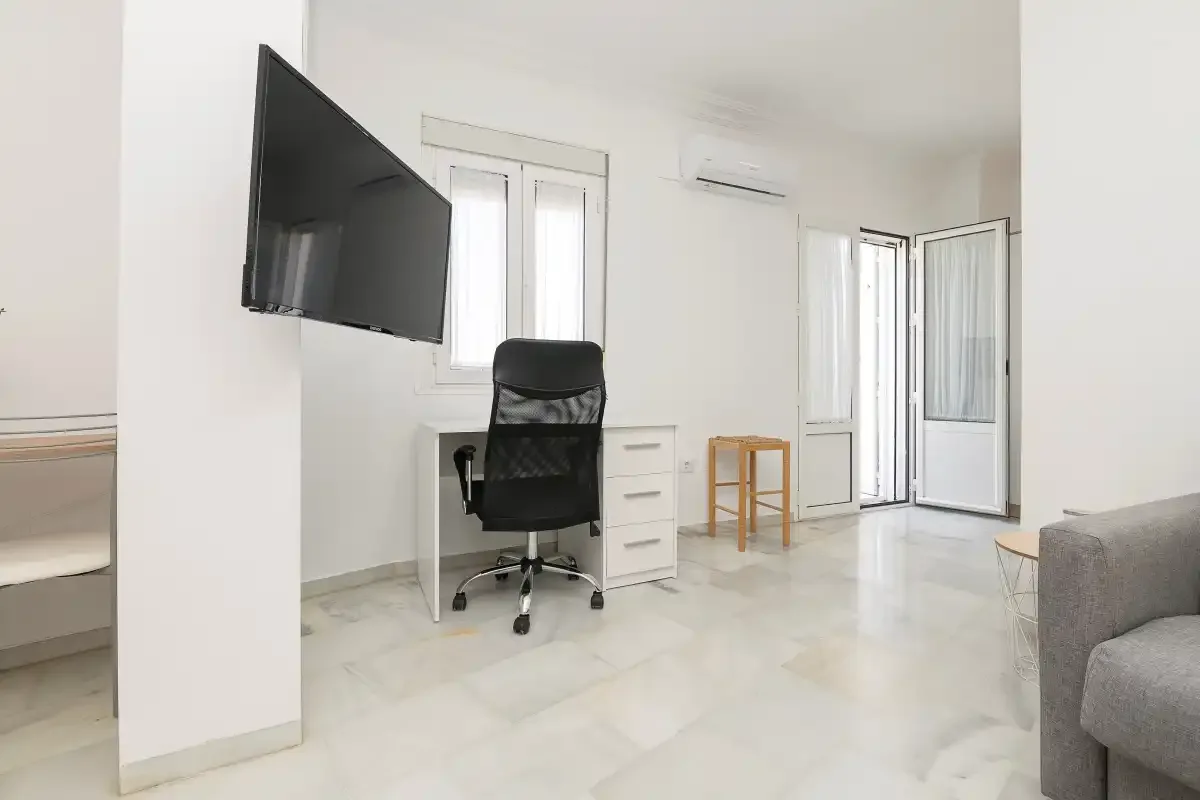 A bright room with a desk, office chair, wall-mounted television, and open balcony doors.
