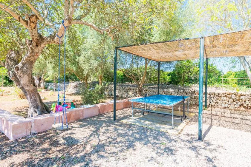Zona de juegos exterior a la sombra, con un columpio y una mesa de ping-pong entre árboles.