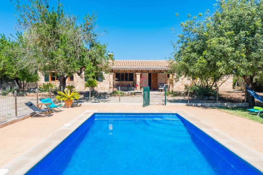 An outdoor swimming pool sits in front of a low house, with trees and lounge chairs nearby.