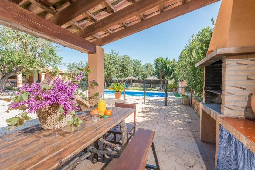 Un patio cubierto tiene una mesa de comedor de madera junto a una barbacoa de piedra y una piscina.