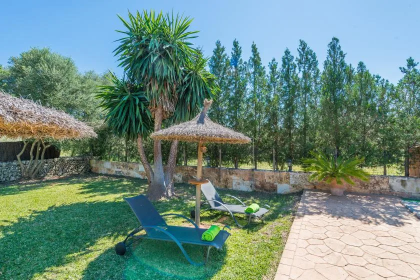 Un patio de jardín soleado tiene asientos reclinables bajo una sombrilla de paja.