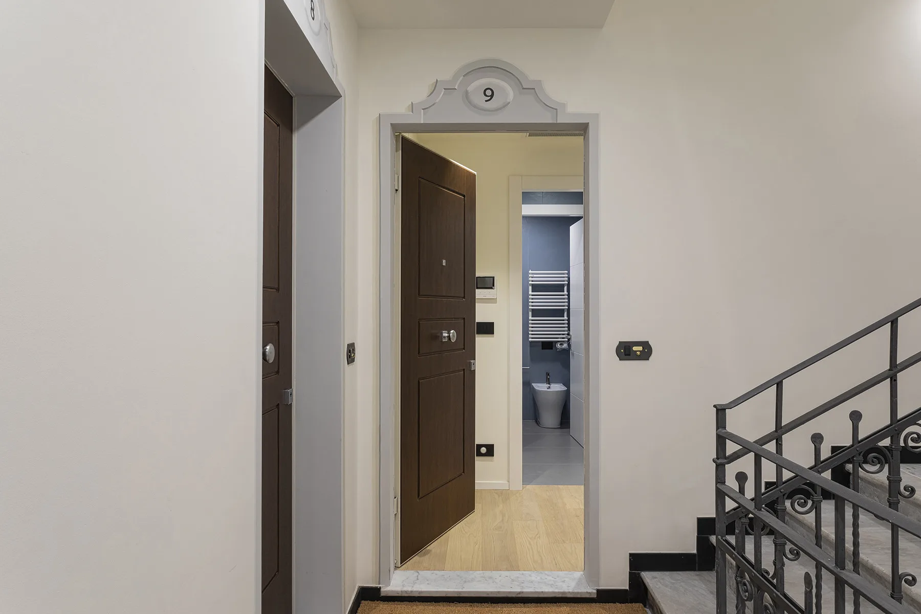 An open entry door reveals a hallway and a bathroom with a toilet and towel warmer.