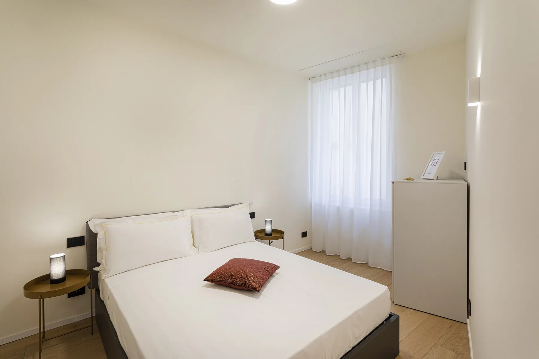 A bedroom with a neatly made bed and a window with sheer curtains.