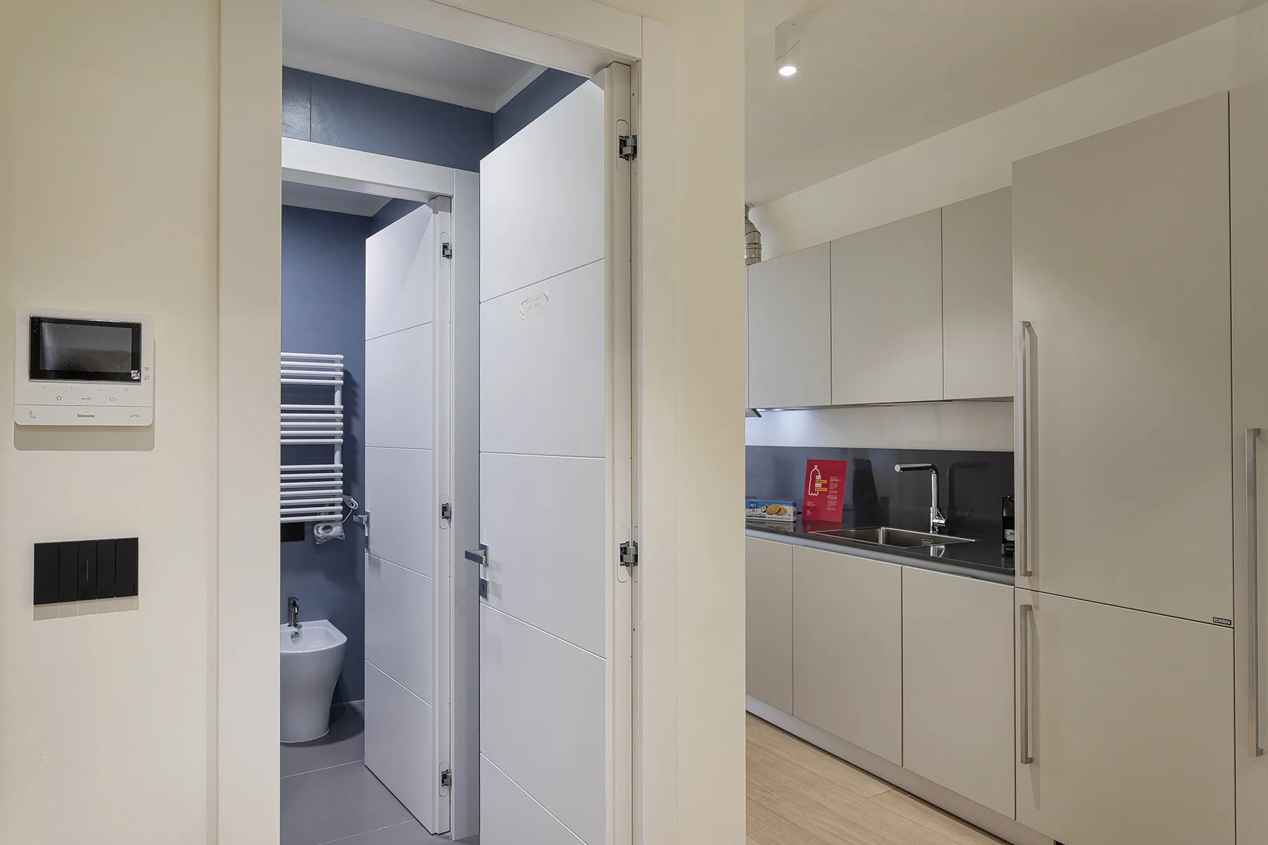 A compact kitchen with pale cabinets is beside a tiled bathroom.